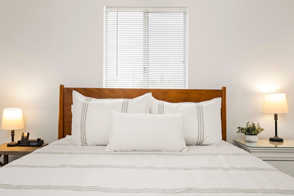 A cozy bedroom features a spacious bed with white linens and subtle striped accents. Soft lighting is provided by bedside lamps on each side, illuminating the room's neutral color scheme. A window with blinds allows natural light to filter in, enhancing the calm ambiance.