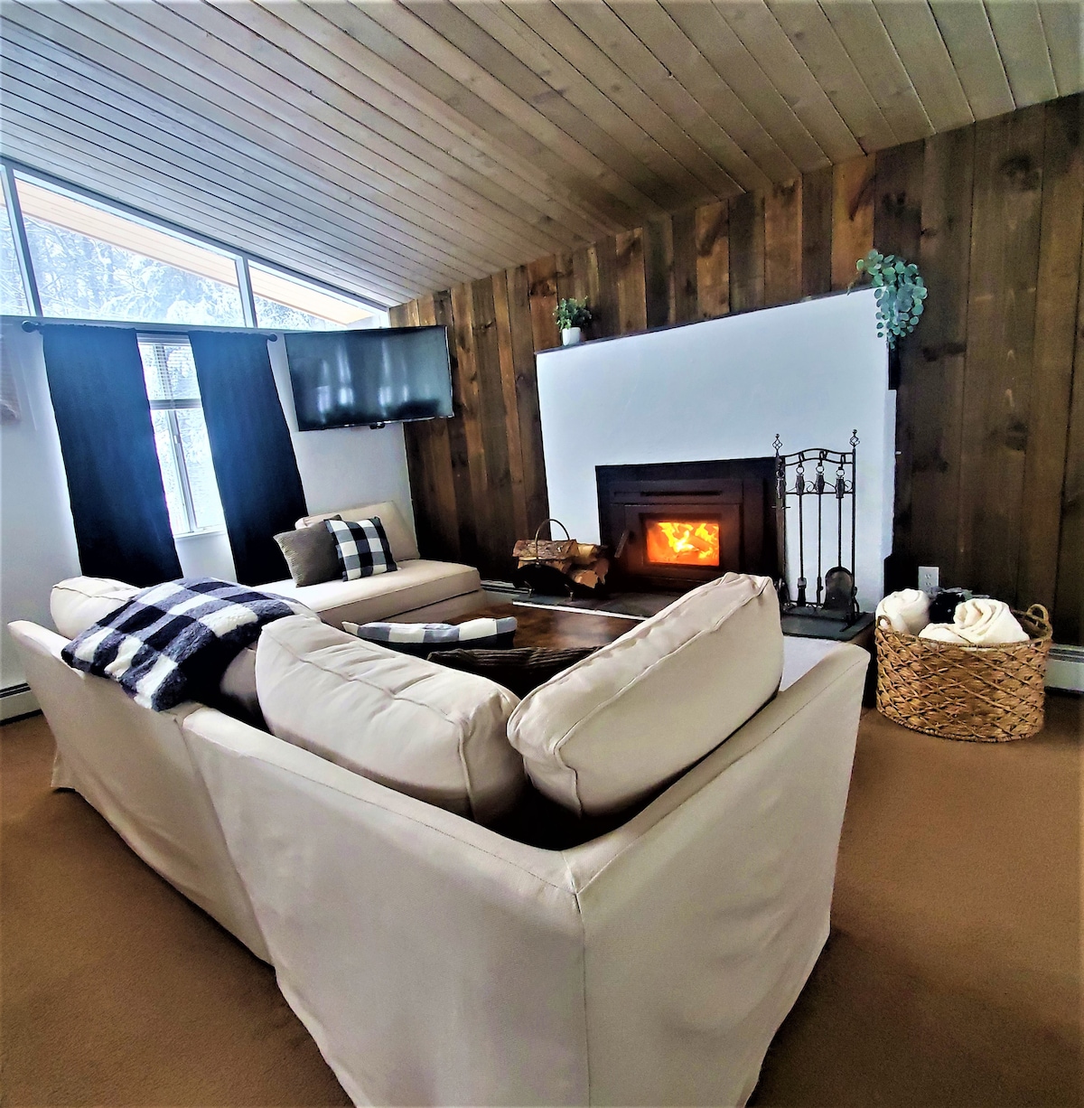 An open living space features a comfortable seating arrangement with light-colored sofas positioned around a wood-burning fireplace. A flat-screen TV is mounted on the wall, and large windows allow natural light to fill the room. Soft textiles and decorative elements enhance the cozy atmosphere.