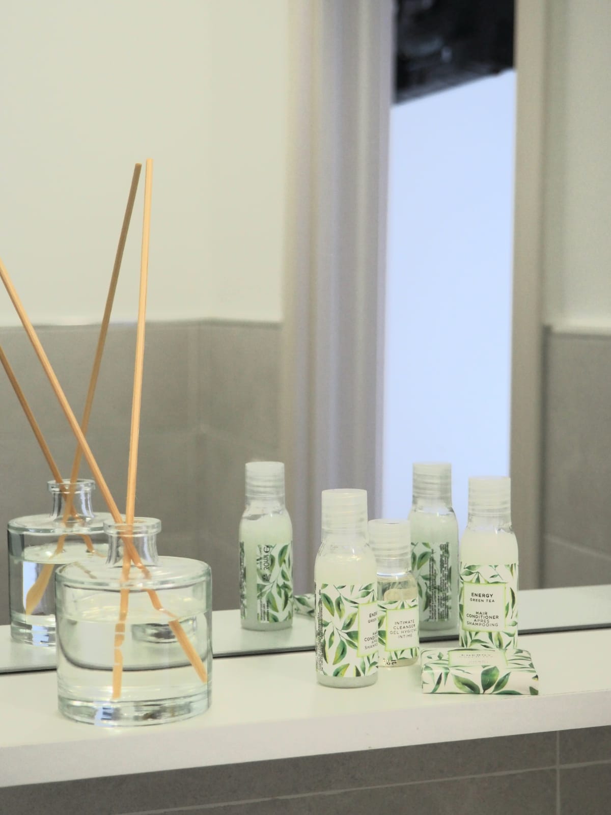 A clean bathroom counter displays a glass vase with reed diffusers and multiple bottles of toiletries, all featuring a botanical design. Natural light reflects softly off the surfaces, enhancing the serene and organized setting.