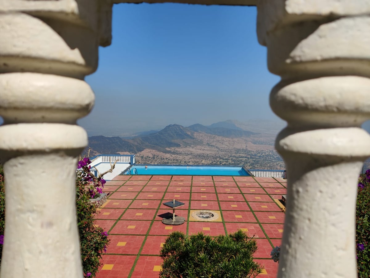 A panoramic view of the private swimming pool is framed by ornate pillars. The vibrant tiled patio displays scattered greenery, while the distant mountains are visible under a clear blue sky. The tranquil pool reflects the stunning landscape surrounding the villa.