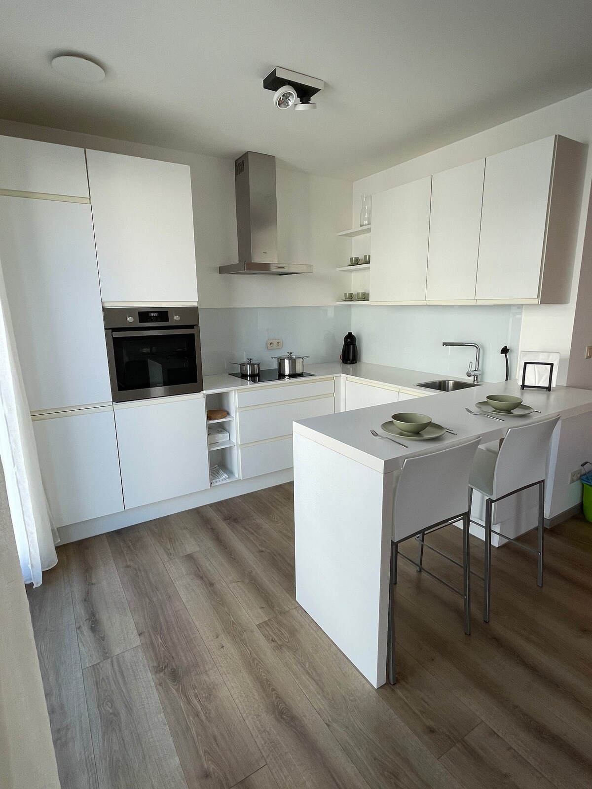 A modern kitchen features sleek white cabinetry and a minimalist design. The countertop includes bar seating for two, complemented by simple dishware. Stainless steel appliances and a glass backsplash reflect natural light, enhancing the bright and clean atmosphere of the space.