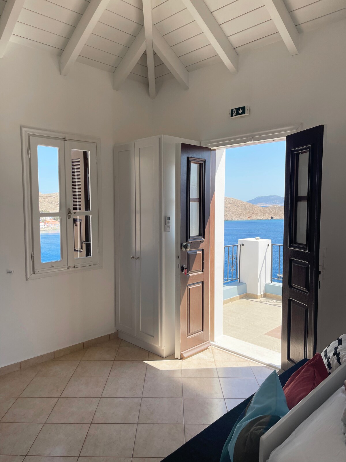 A bright and airy entrance showcases two dark wooden doors, open to reveal a view of the blue sea beyond. Natural light fills the space, accentuating the light-colored walls and tiled floor, while a cozy bed is partially visible in the foreground.