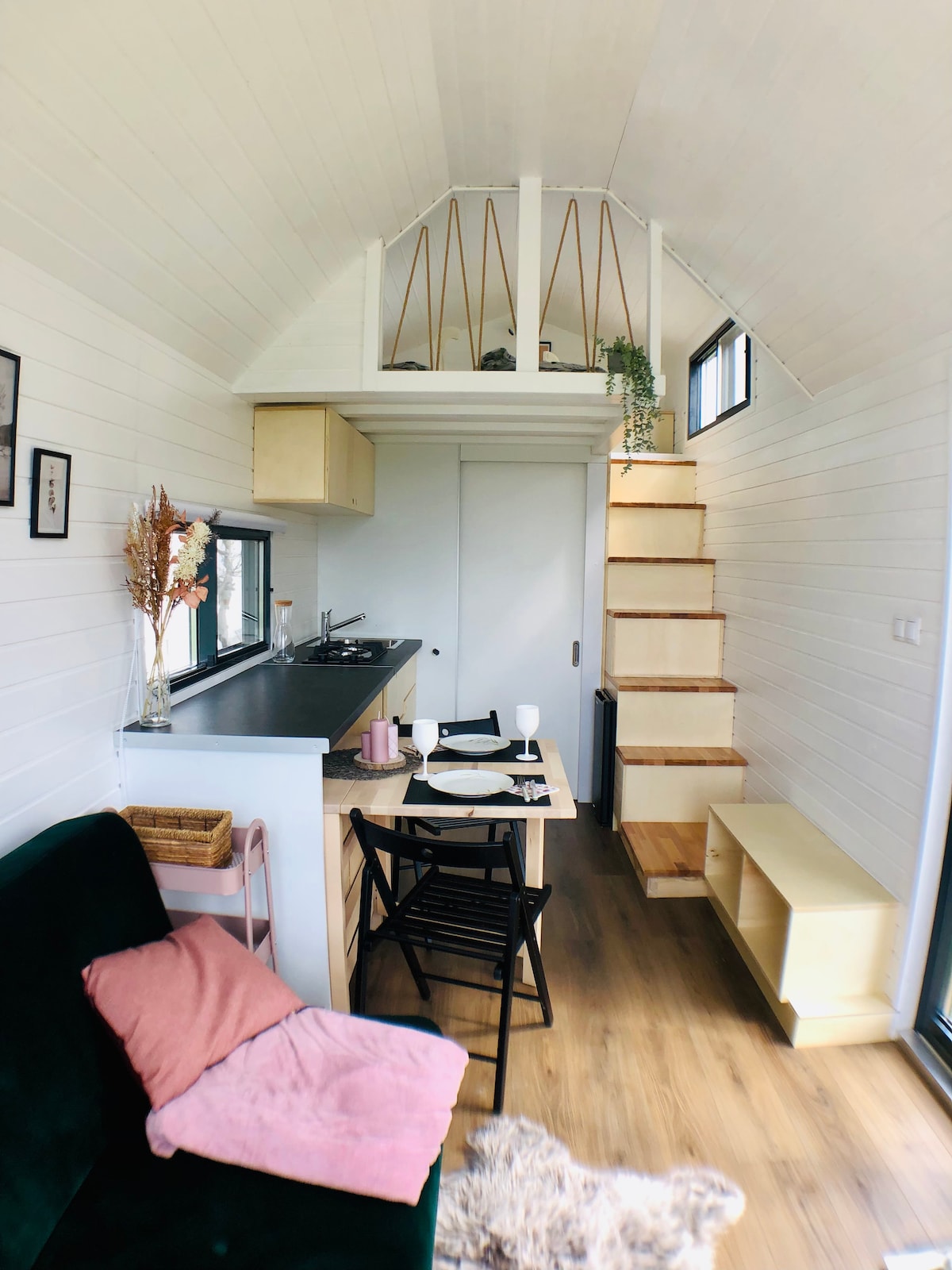 An inviting interior of a tiny house features a compact kitchen along one wall, a dining area with a small table set for two, and a staircase leading to a lofted sleeping area. Natural light floods in through large windows, enhancing the airy atmosphere.