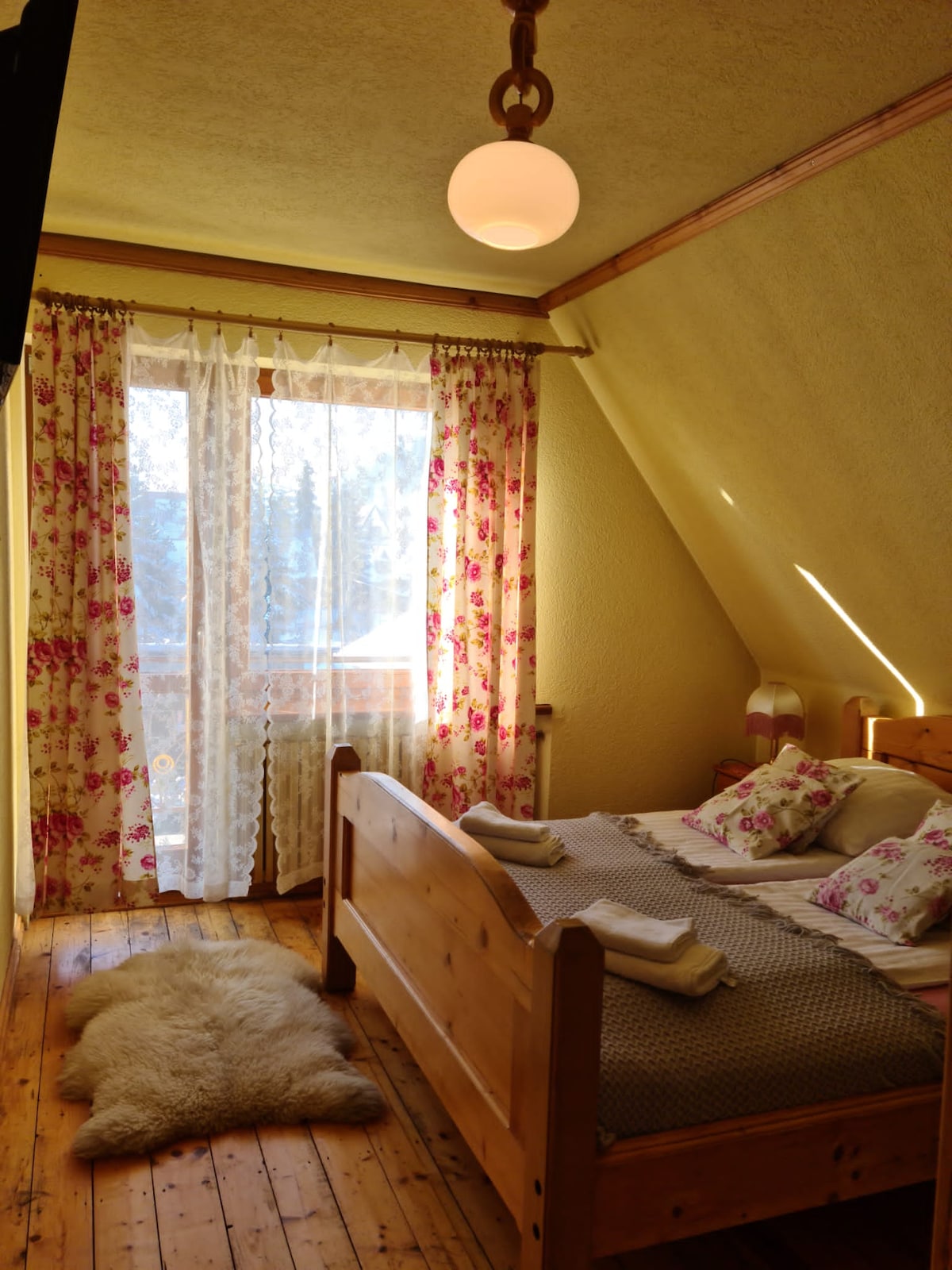 A cozy bedroom features a wooden bed adorned with floral-patterned pillows. Natural light enters through a floral-curtained door leading to a balcony. A soft rug lies beside the bed, and the sloped ceiling adds character to the space.