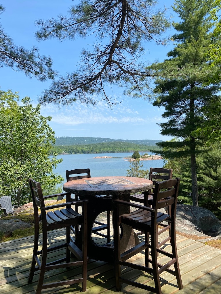 An outdoor seating area features a round table surrounded by four high-backed chairs, set on a wooden deck. Lush trees and a clear view of the water and distant landscape create a peaceful setting for relaxation.