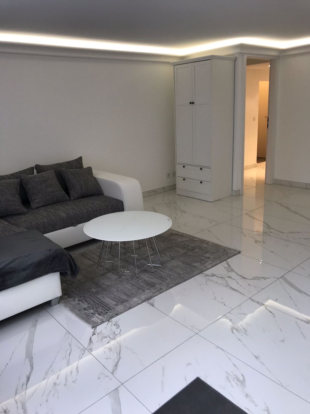 A modern living area is highlighted by a sleek, white sofa adorned with gray cushions. A small round table is positioned on a textured rug over polished marble flooring. The space is complemented by neutral-colored walls and soft lighting, creating an airy ambiance.