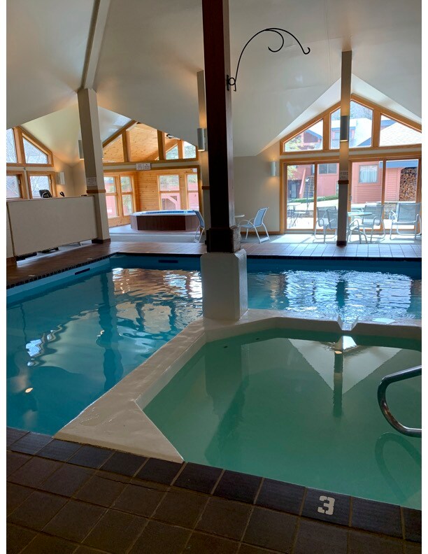 An indoor pool area showcases a blend of refreshing blue water and inviting light. Large triangular windows allow natural light to fill the space, while a hot tub is visible in the corner. Lounge chairs are positioned nearby, promoting relaxation.