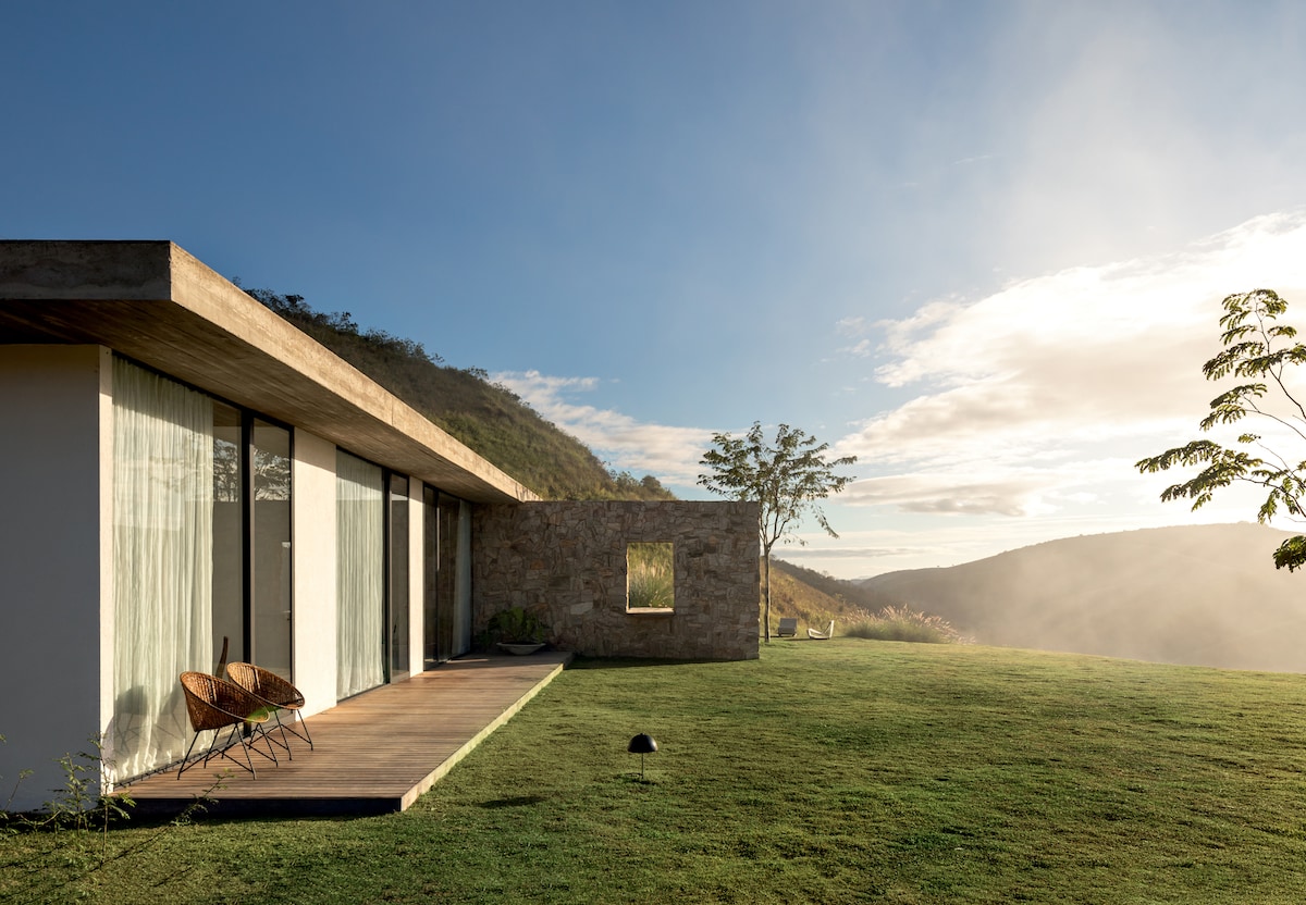 The exterior of a modern home is shown, featuring expansive glass panels and a concrete overhang. A stone wall adds texture alongside a landscaped lawn. Two chairs are positioned on the wooden deck, offering a view of the surrounding natural landscape under a soft, early morning light.