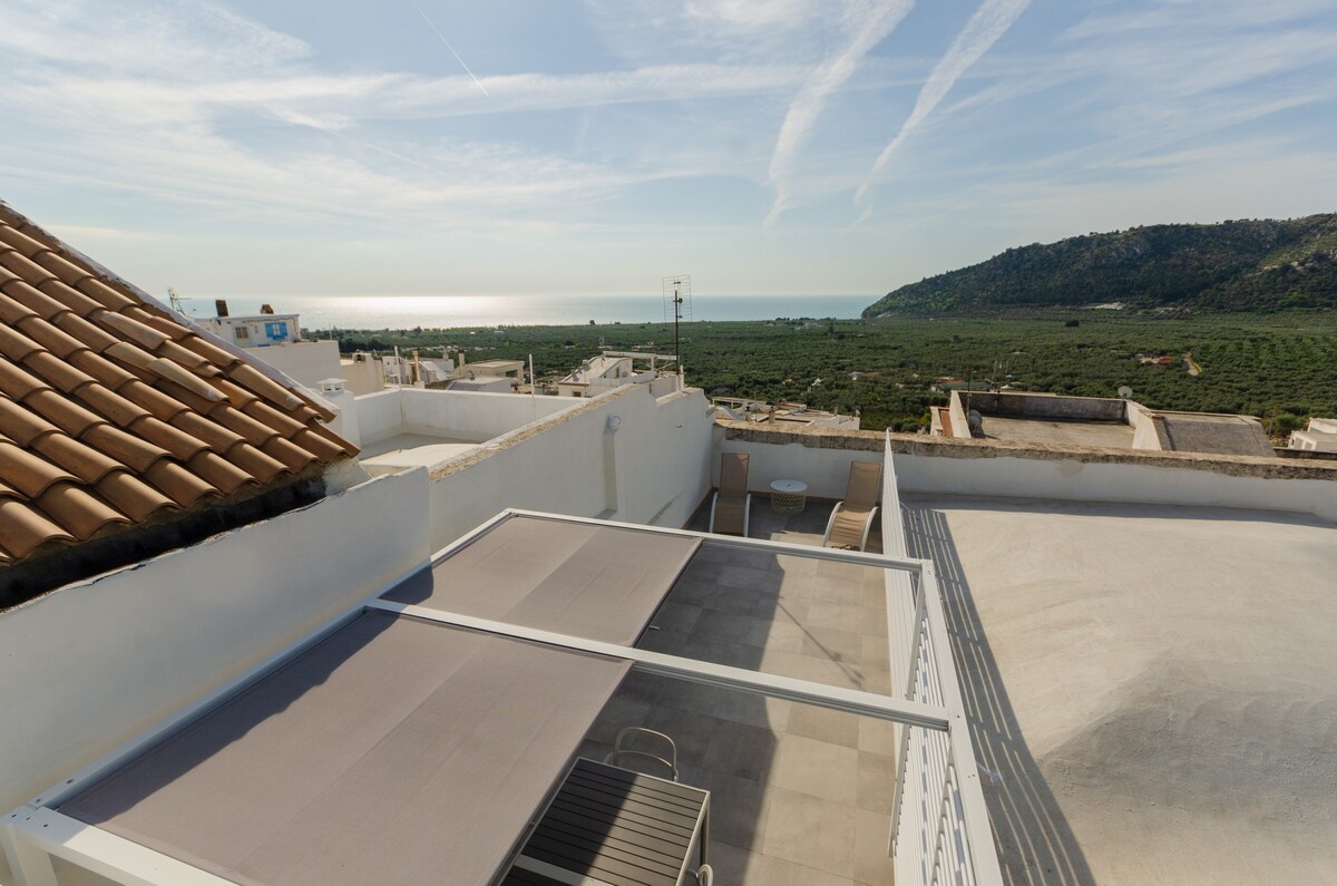 A panoramic terrace offers views of the sea and surrounding olive groves. The roof features a sun canopy and seating area. The landscape includes distant hills and an expansive coastline under a clear blue sky with wispy clouds.
