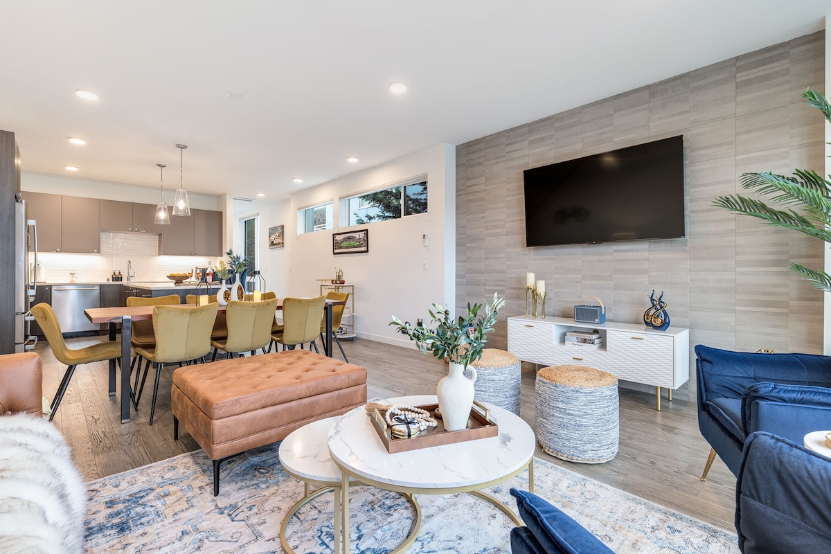 A modern living space features an open layout with a sleek kitchen and dining area. Comfortable seating is arranged around a stylish coffee table. Natural light floods through large windows, illuminating the neutral color palette and contemporary furnishings. A flat-screen TV is mounted on the wall.