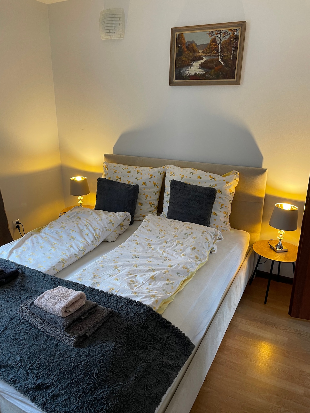 A cozy sleeping area features a spacious bed with a light-colored duvet and two decorative pillows. Soft bedside lamps provide warm illumination, and a picture hangs above the bed. A textured blanket and rolled towels rest on the bed's edge, offering welcoming touches.