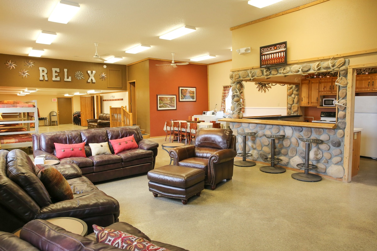 The spacious dining and living area is furnished with multiple leather sofas, complemented by a matching armchair. A stone bar with high stools is positioned against the wall. The room features warm colors and a welcoming atmosphere for group gatherings.