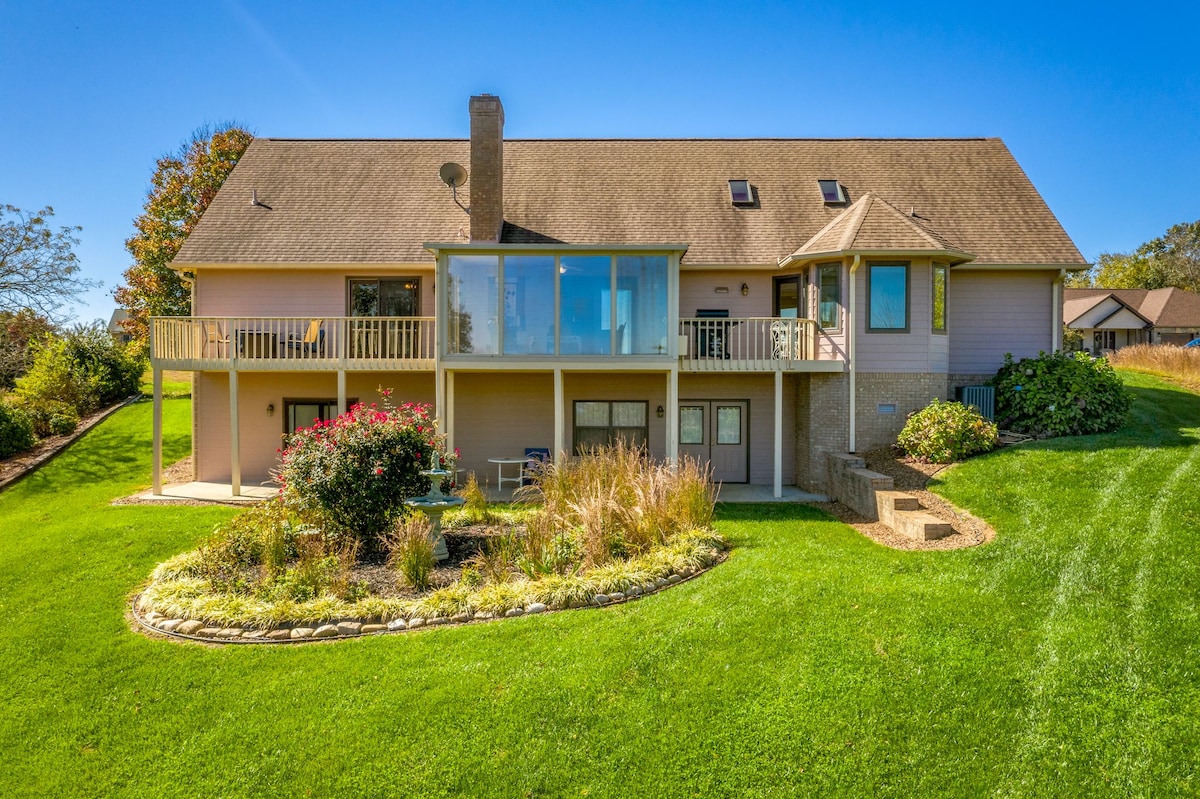 The exterior of a two-story home is surrounded by well-maintained lawns and colorful landscaping. A spacious wraparound porch with seating overlooks the yard. Large windows enhance natural light, and a pathway leads to the garden area, creating an inviting space for outdoor enjoyment.