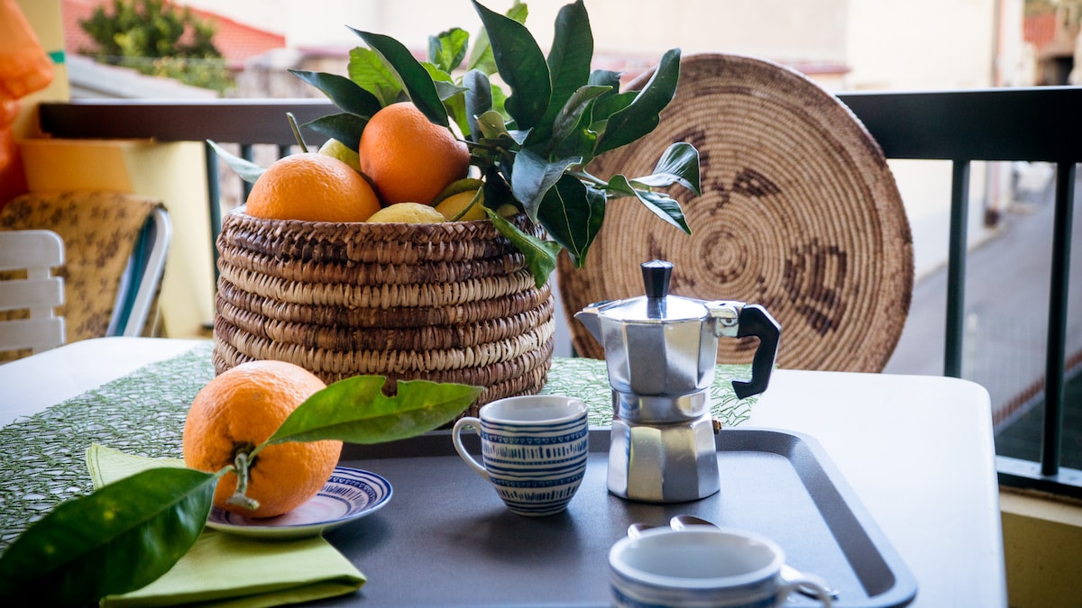 A balcony scene features a wicker basket filled with fresh oranges and lemons. A small coffee pot and two cups are placed on a tray atop a table. The backdrop includes a woven decorative piece, enhancing the outdoor dining experience.