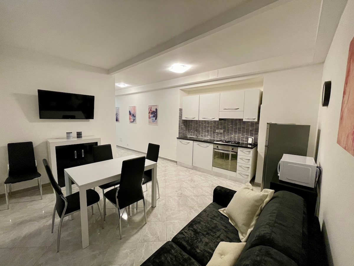 An open-plan living area is depicted, featuring a modern kitchen with sleek cabinetry and appliances. A dining table with six chairs is situated beside the kitchen, and a dark sofa is positioned in the foreground. Bright artwork adorns the walls, enhancing the spacious ambiance.
