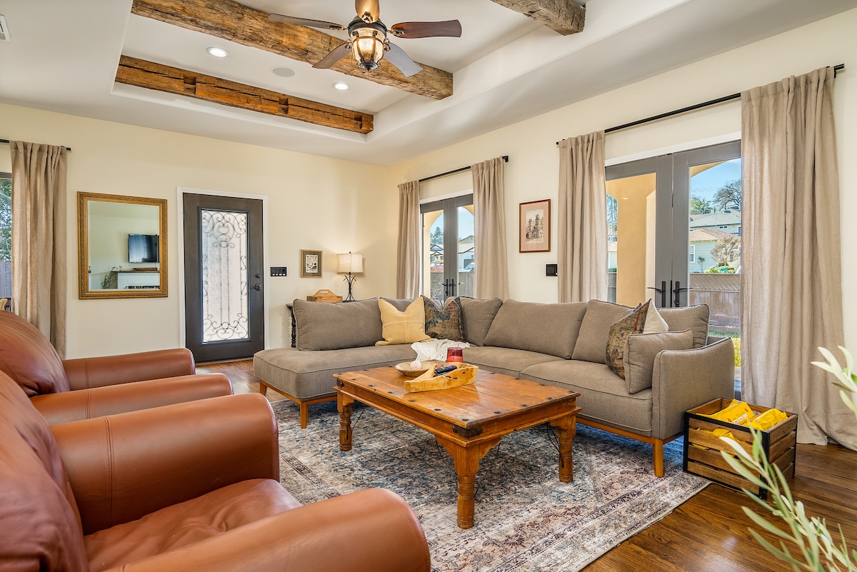 A spacious living room features a comfy sectional sofa alongside two leather club chairs. Natural light floods the area through large doors leading to the outdoor space. Exposed wooden beams and soft curtains add a cozy touch to the inviting atmosphere.