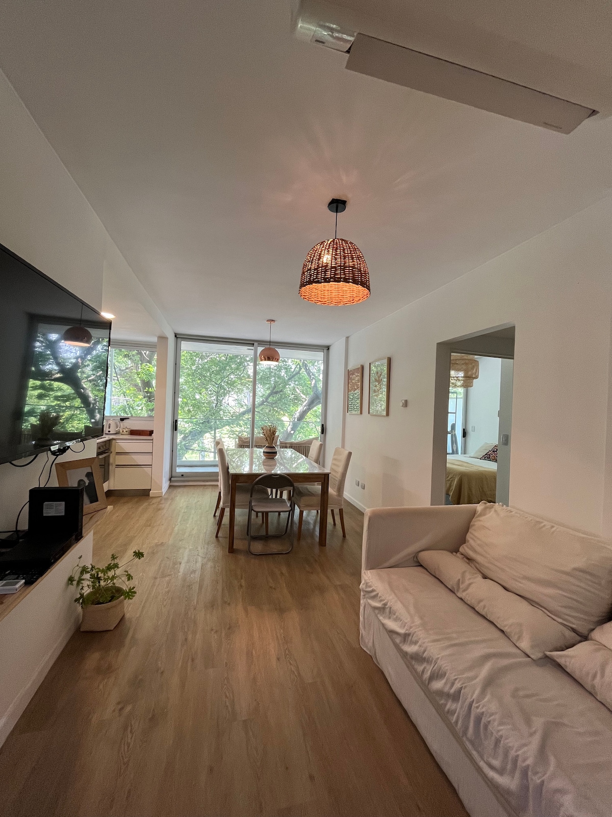 A modern living area is highlighted by a cozy sofa facing a wall-mounted television. A dining table with chairs is positioned by large windows that allow natural light to fill the space. Decorative pendant lights hang from the ceiling, enhancing the room's ambiance.