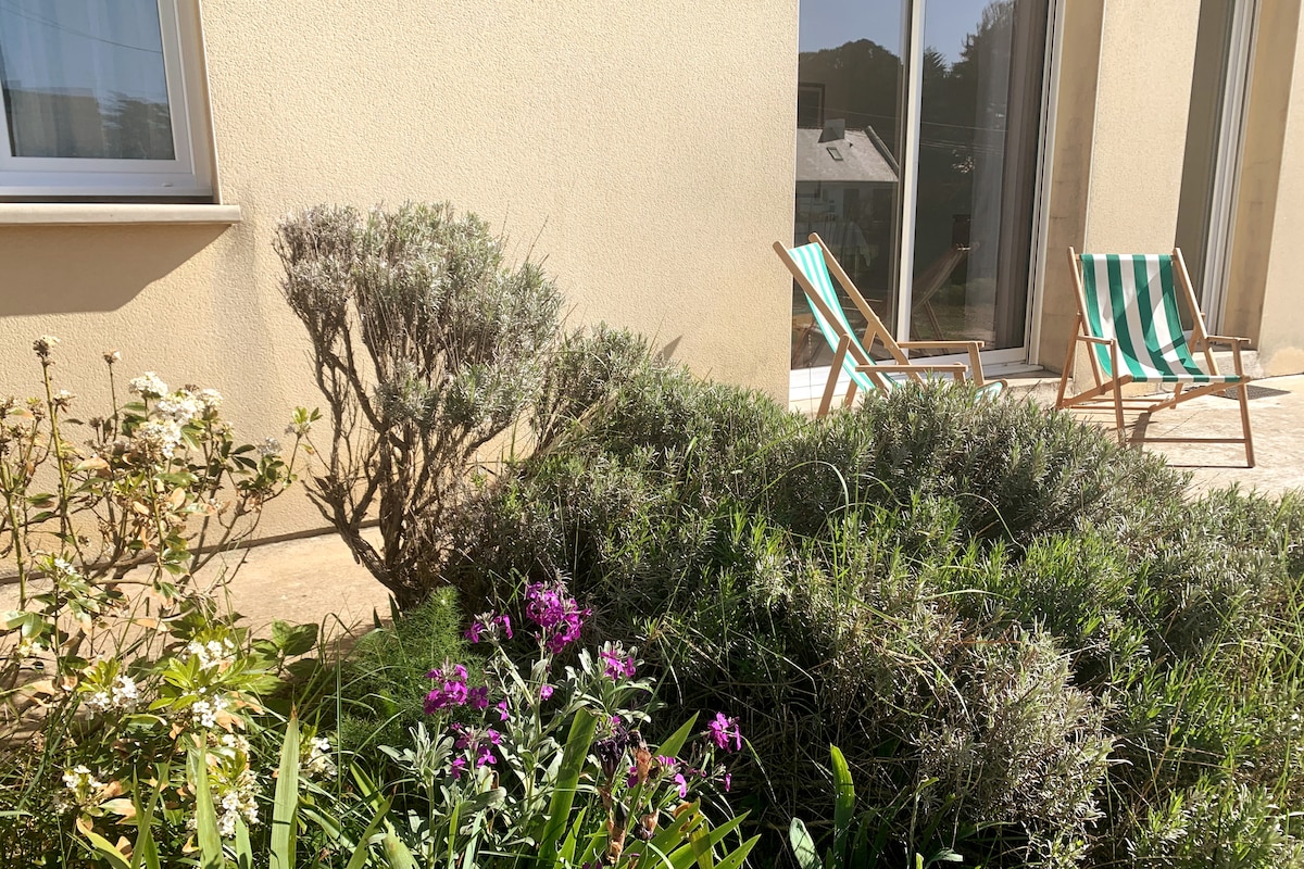 A garden area is visible in this image, featuring lush green foliage and colorful flowers, including purple blooms. Two striped deck chairs are positioned on a stone patio, providing a place to relax outdoors. Sunlight highlights the greenery, creating a serene atmosphere.