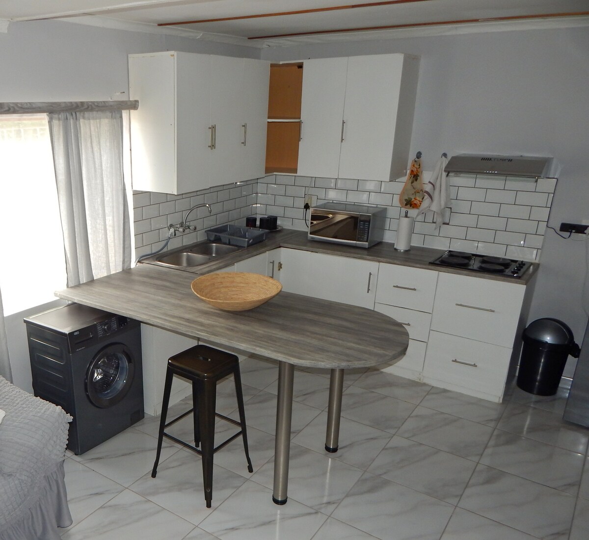 A modern kitchen space is showcased, featuring white cabinetry and a laminate countertop. Stainless steel appliances include a stove and microwave, alongside an integrated washing machine. Natural light enters through a window, highlighting a round wooden bowl placed centrally on the kitchen island.