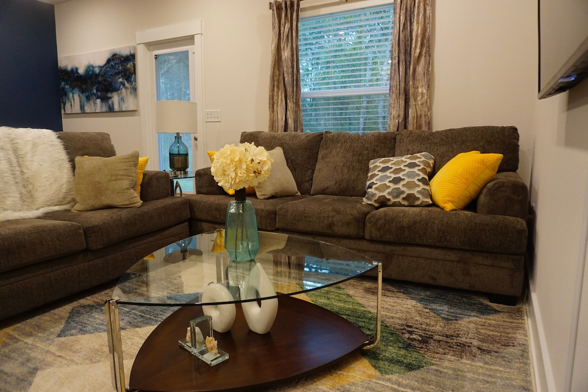 A spacious living room features a large sectional sofa adorned with various cushions in soft tones. A glass coffee table sits in the center, displaying decorative items. Natural light filters through a window, complemented by sheer curtains, while a piece of art hangs on the adjacent wall.