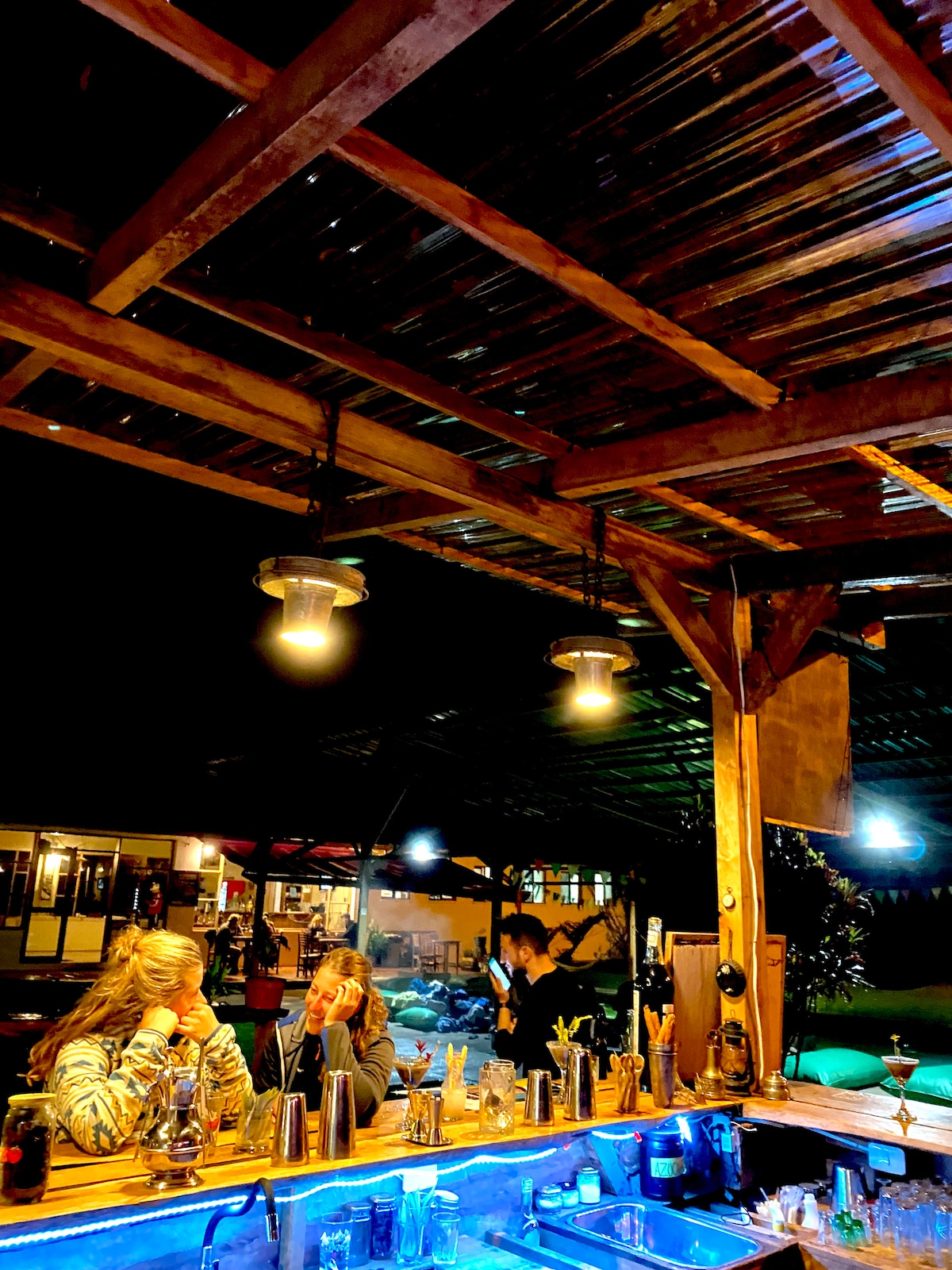 The bar area features a wooden counter, illuminated by warm pendant lights hanging from a corrugated roof. Behind the bar, various bottles and utensils are neatly arranged. Guests are seated, enjoying their drinks in a lively atmosphere, with soft lighting enhancing the surroundings.
