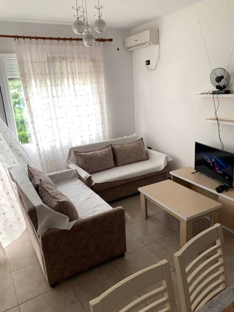 A light-filled living area features two sofas arranged around a small table. A television is positioned on a shelf across from the seating, with soft curtains allowing natural light to enter from a nearby window.