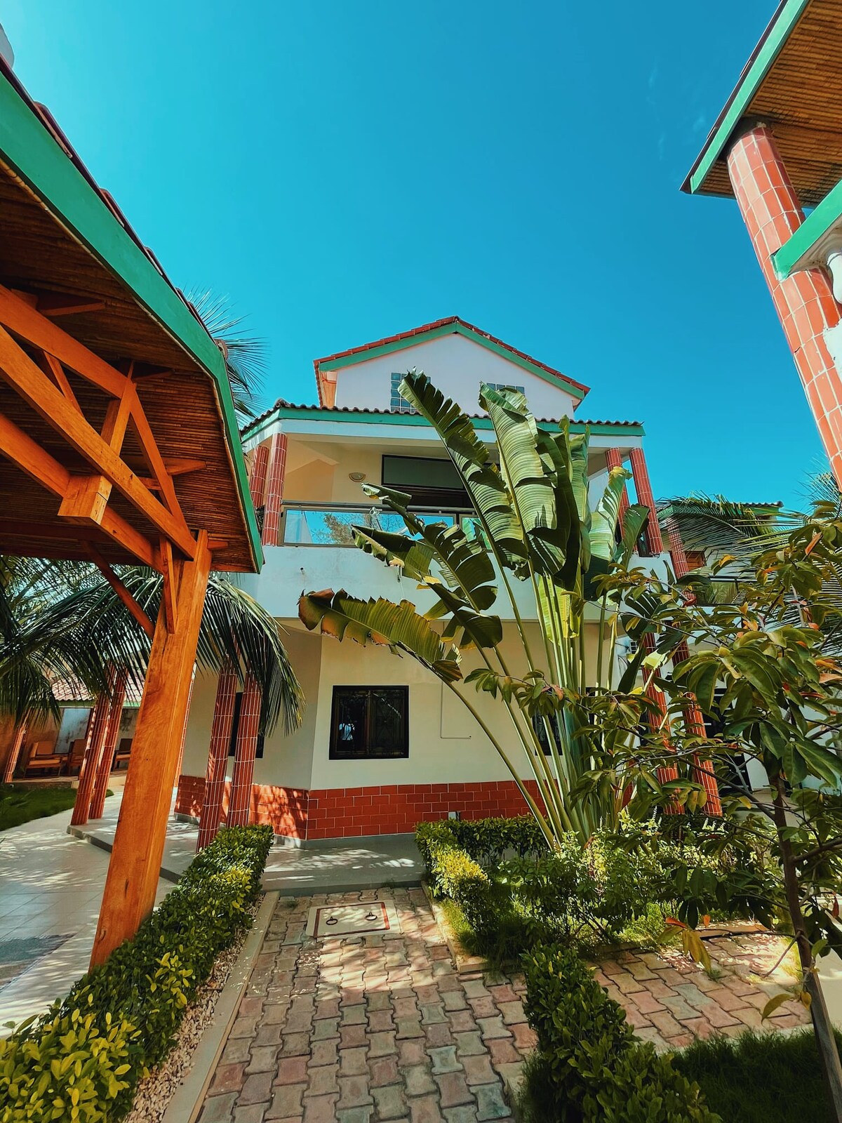 A multi-story house with a vibrant white and terracotta façade is framed by lush greenery, including tall palm leaves. The structure features large windows that invite light, while a pathway lined with manicured hedges adds to the outdoor ambiance.