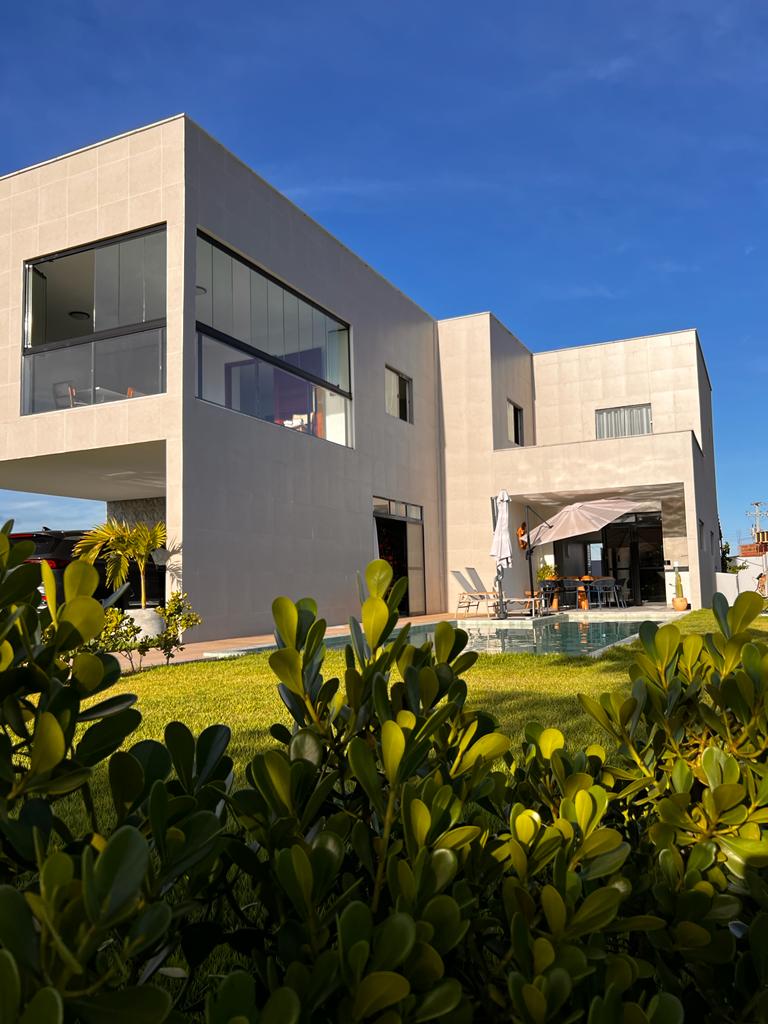 The modern exterior of the house showcases large glass windows, allowing abundant natural light. A lush green lawn surrounds a private pool area, complete with outdoor furniture tucked under a shade structure. The building design emphasizes clean lines and a contemporary aesthetic.