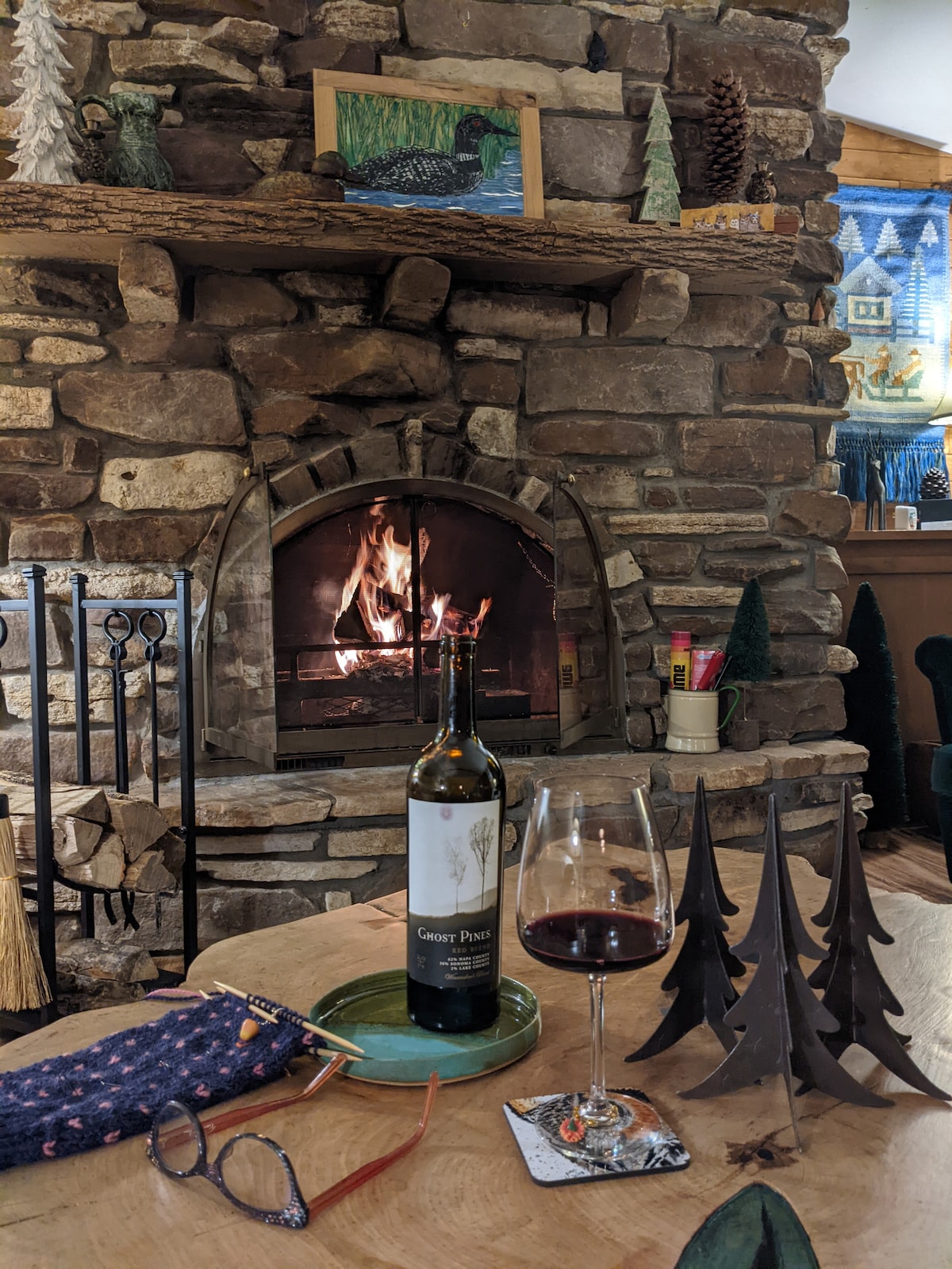 A warm stone fireplace serves as a focal point, with a gently flickering fire visible inside. A bottle of wine and a glass sit on a wooden table, accompanied by a small plate. Decorative trees are arranged nearby, enhancing the cozy ambiance.