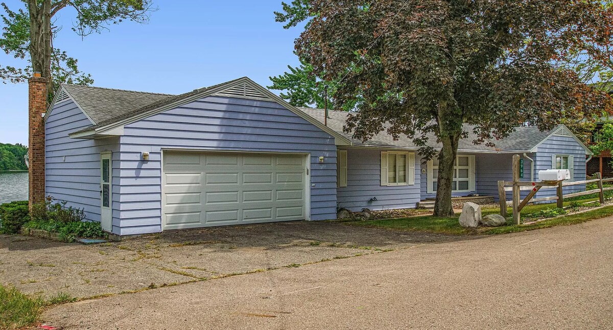 A charming single-story house is set against a tranquil lake backdrop. The exterior features light blue siding and a spacious driveway leading to a two-car garage. Lush greenery and trees line the property, enhancing the serene surroundings.
