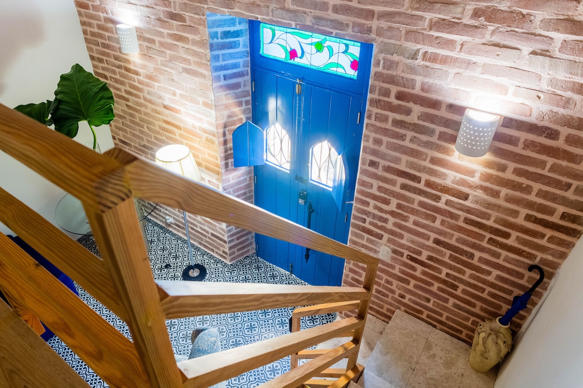 A bright entrance area is presented with a cozy staircase leading down, featuring wooden steps and a stylish railing. A vibrant blue door with decorative glass is centered, surrounded by warm brick walls, while patterned tiles add a touch of character to the floor.