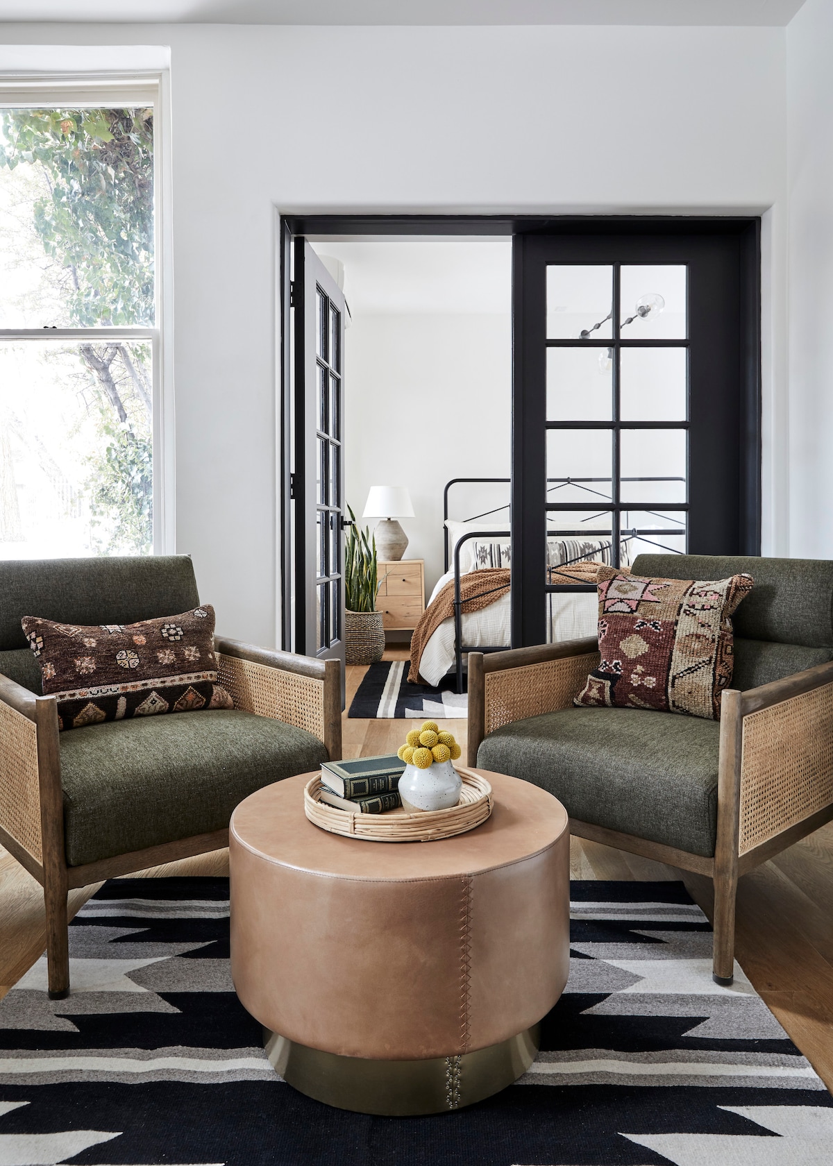 An inviting seating area features two stylish chairs with earthy tones, positioned beside a round leather ottoman. Natural light filters through a nearby window, illuminating the space. Elegant French doors lead to the bedroom, enhancing the open layout.
