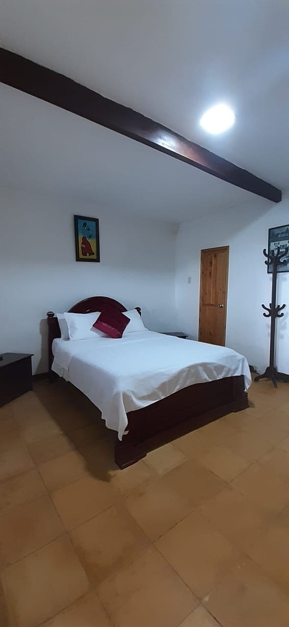 A cozy sleeping area features a wooden bed dressed in white linens, complemented by a red throw pillow. A small bedside table is visible next to the bed, and a colorful piece of wall art adorns the room. A door leads to an adjacent space.