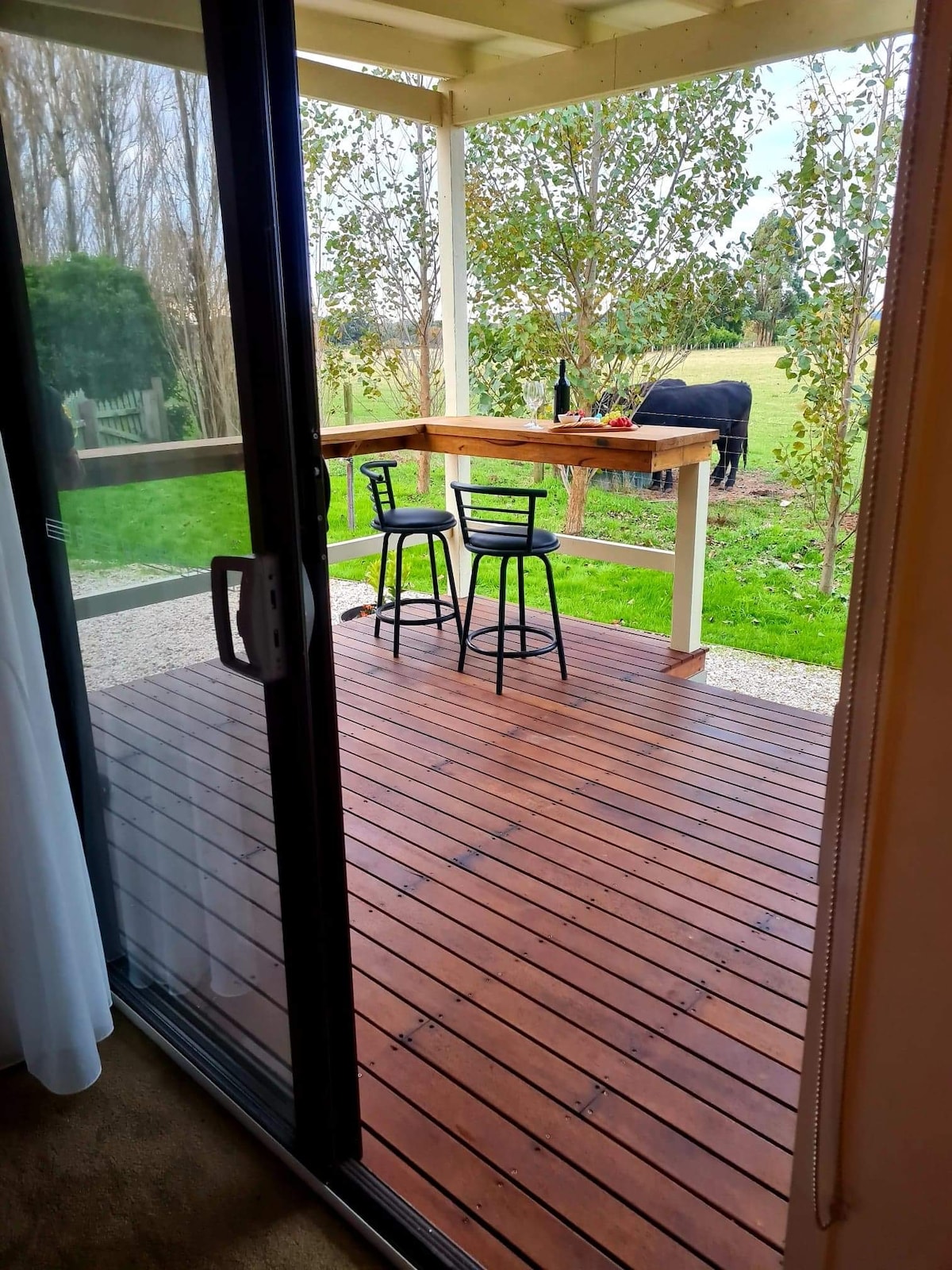 A wooden deck is highlighted, featuring a bar table with two black chairs. The area is framed by lush greenery and overlooks a grassy field, where a black animal can be seen grazing. Large glass doors provide an inviting entryway.