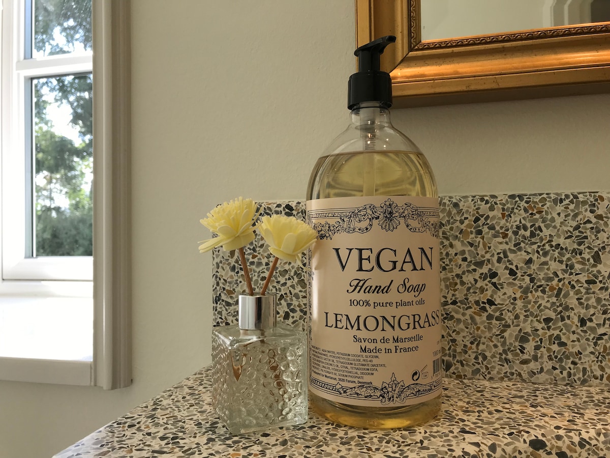 A large bottle of vegan hand soap with a lemongrass scent is displayed on a textured countertop. A small, glass vase containing delicate flowers adds a touch of decoration. Natural light streams in through a nearby window, enhancing the clean and tidy environment.