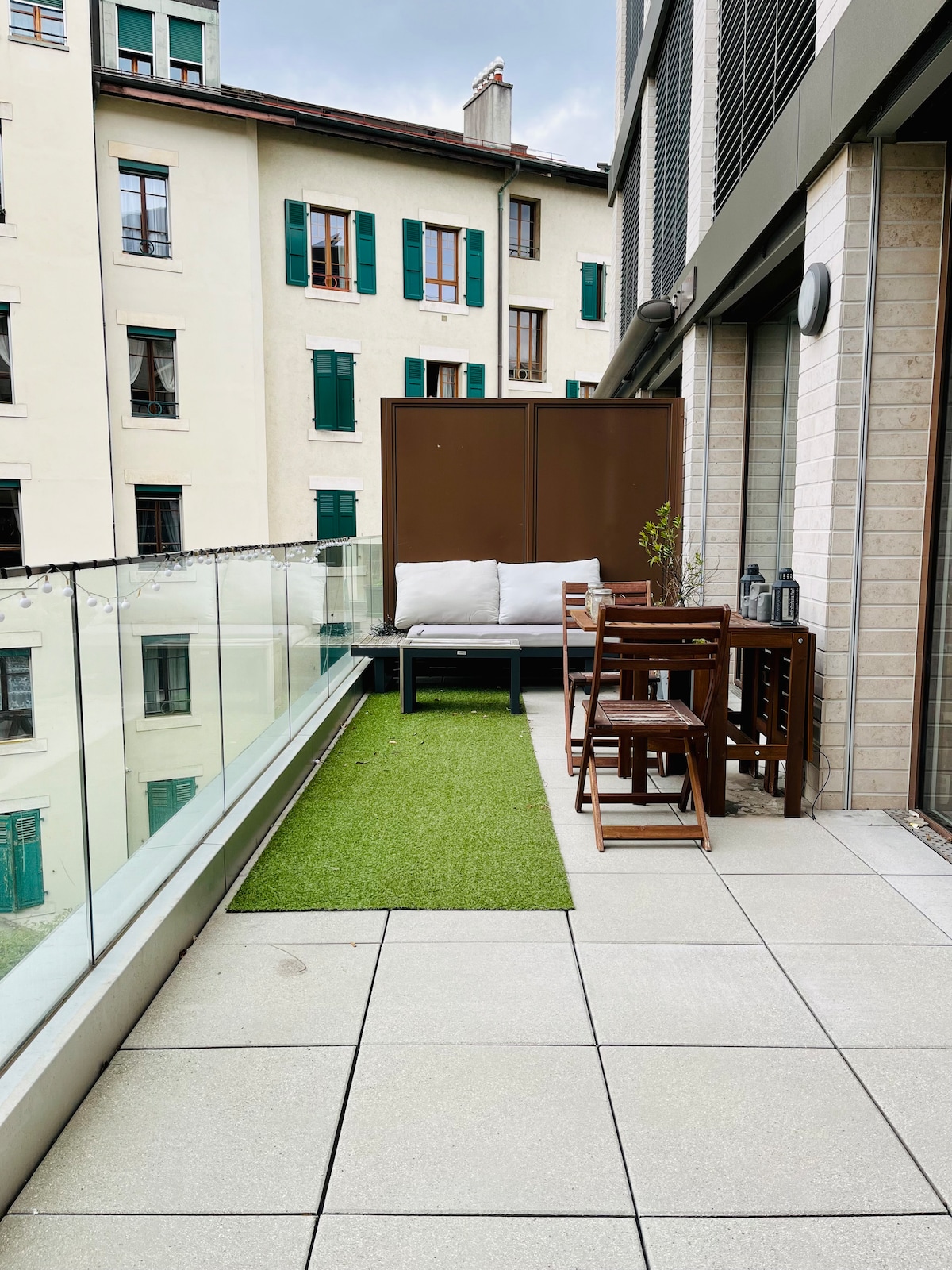 A terrace is featured with contemporary outdoor furniture, including a seating area with cushions and a small dining table. Green grass-like turf is laid out, and glass railings provide an unobstructed view of the surrounding buildings.