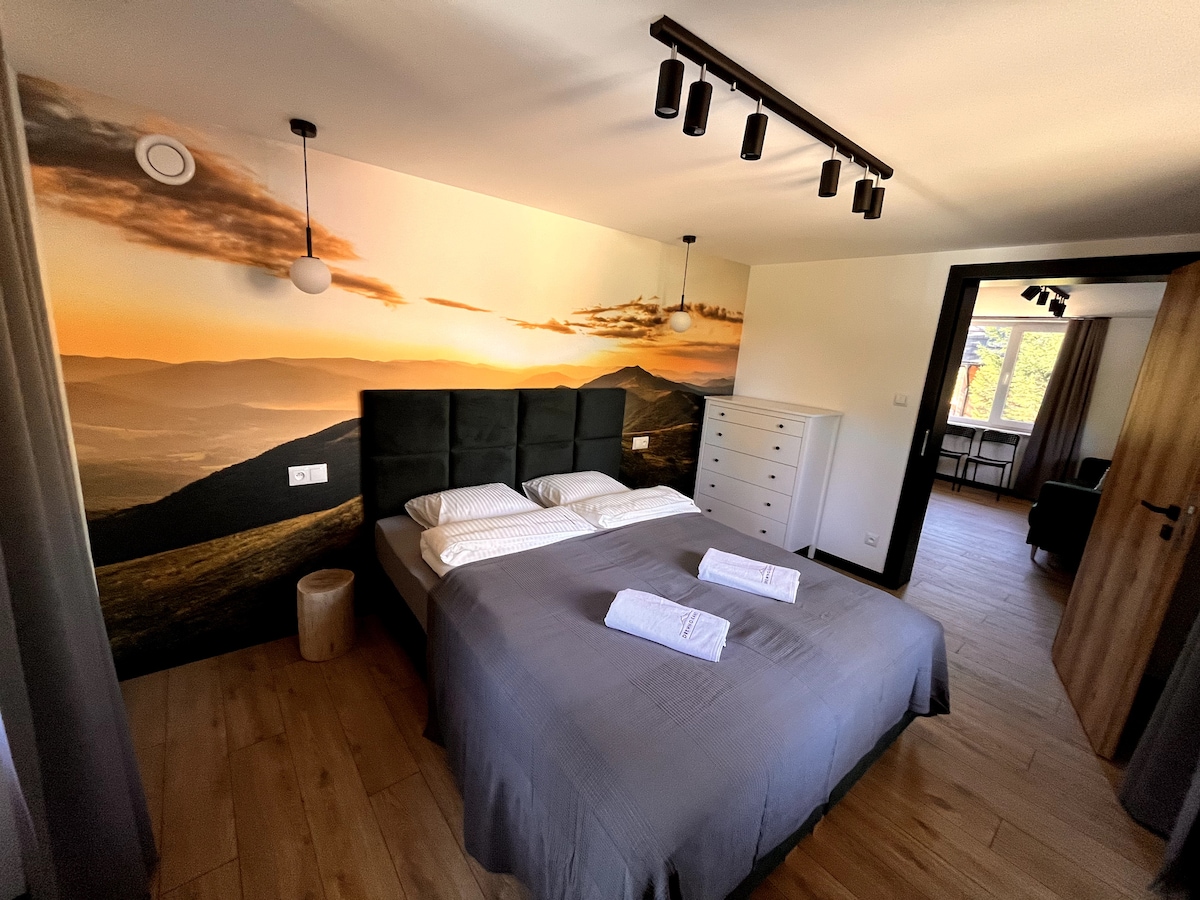 A well-lit bedroom features a spacious bed with neatly arranged linens and towels. A large wall mural displays a scenic sunset over mountains, complementing the warm tones of the wooden floor. Modern lighting fixtures are suspended from the ceiling, enhancing the overall atmosphere.