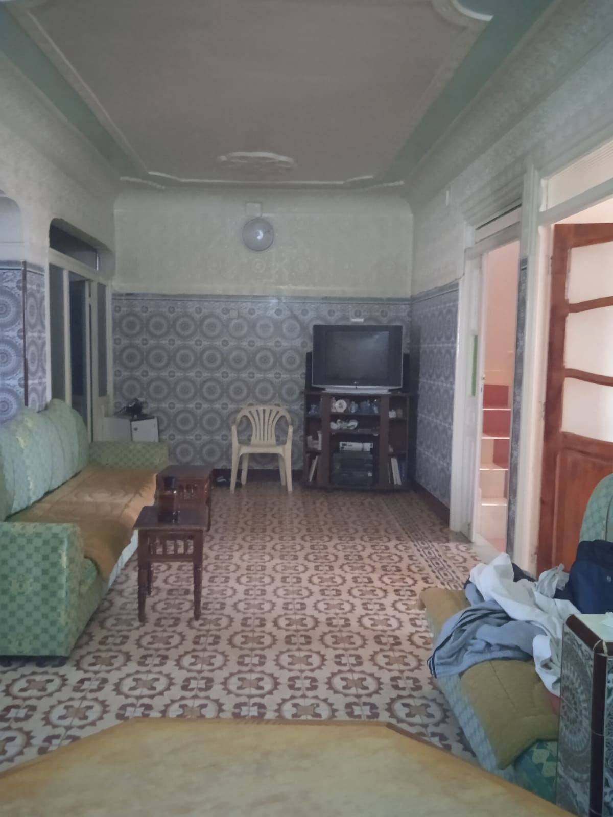 A living area features patterned tile flooring and ornate wall designs. Two green sofas are positioned around a small wooden coffee table. A TV is situated on a stand against the wall. A chair is located in the corner, with light streaming in through the windows.