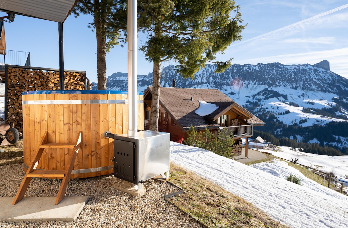 A wooden hotpot is situated on a gravel area, accompanied by a metal stove. Pine trees frame the scene, with snow-covered ground extending towards a backdrop of mountains. A chalet is visible in the distance under a clear blue sky.