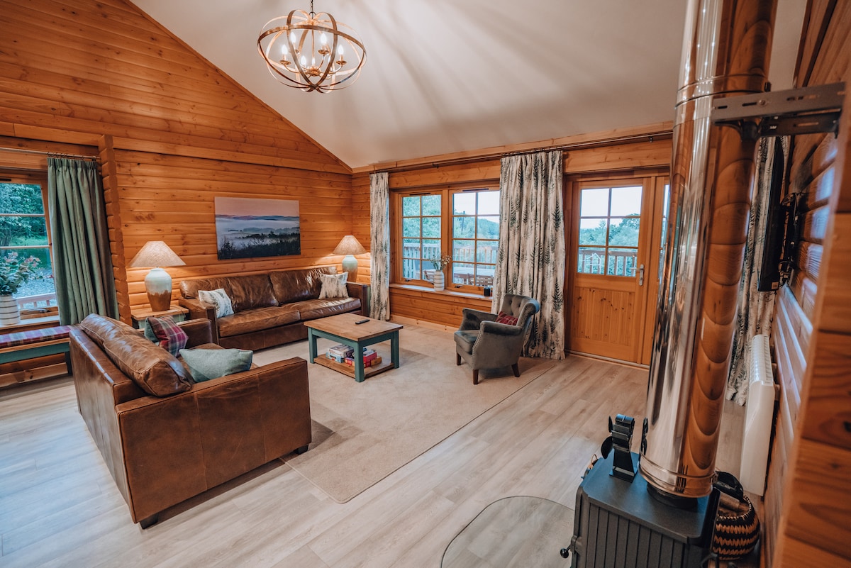 The spacious living area features a high ceiling and warm wooden paneling. A comfortable leather sofa and armchair are arranged around a coffee table. Large windows allow natural light and offer views of the surrounding woodland. A log-burning stove adds a cozy touch.