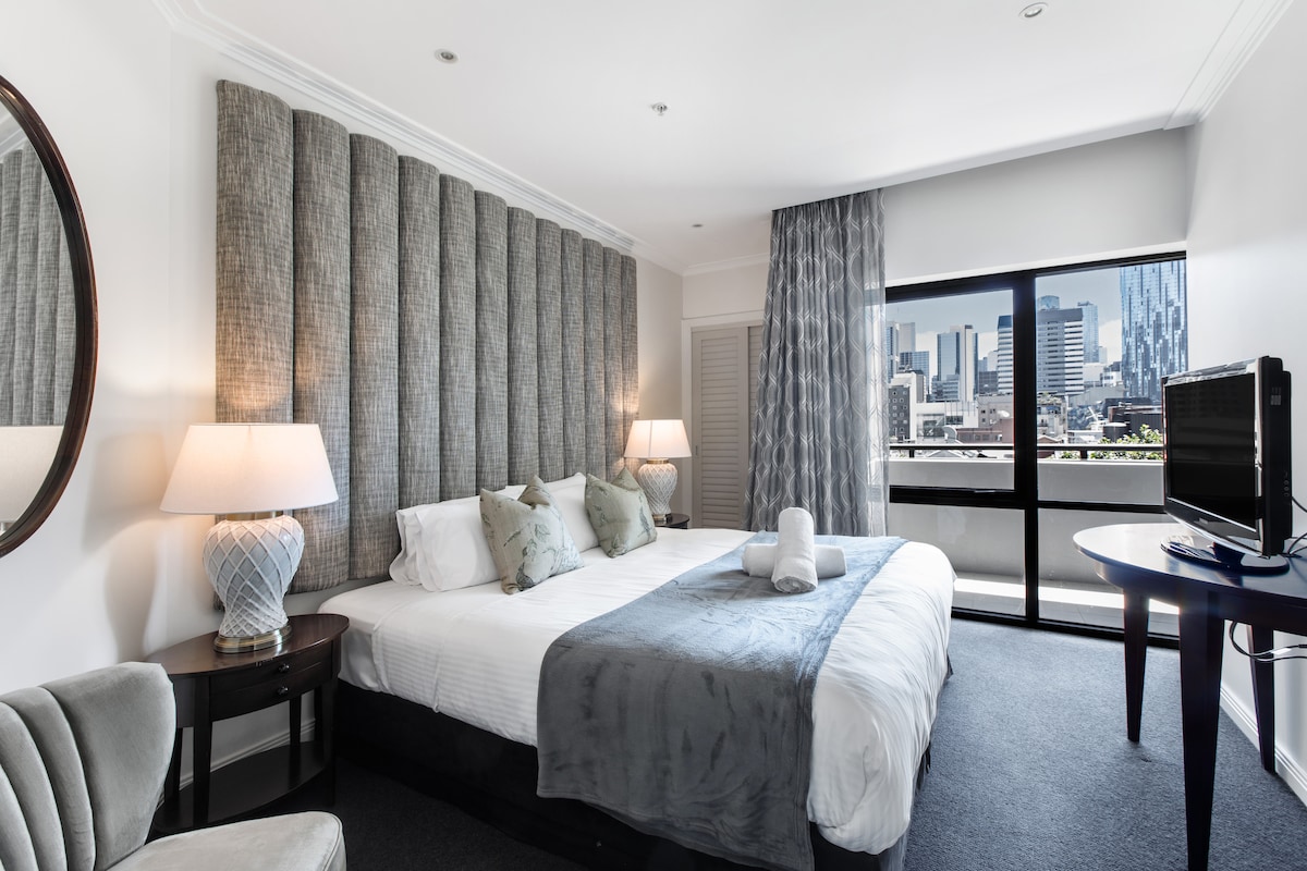 A spacious bedroom features a large bed adorned with plush pillows and a cozy blanket. Floor-to-ceiling windows offer sweeping views of the city skyline. A contemporary armchair and circular table complete the space, with soft lighting provided by two bedside lamps.