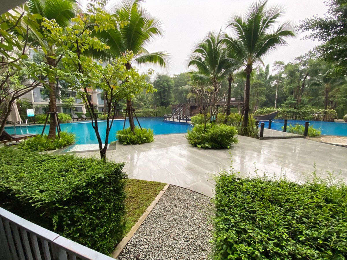 A serene courtyard area features well-maintained greenery and smooth stone pathways. The central focus is a large swimming pool surrounded by palm trees, providing a sense of tranquility. The distant view of additional pool features can be seen amidst the lush landscape.