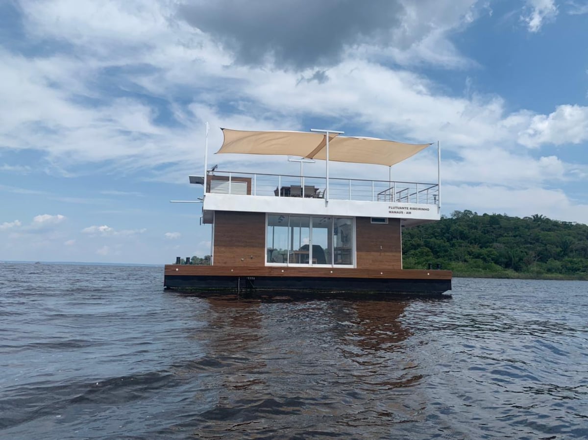 A floating eco-friendly structure is presented on the water, featuring an upper deck with shade provided by a sail. Large windows offer views of the surroundings, while natural wood elements enhance the modern design.
