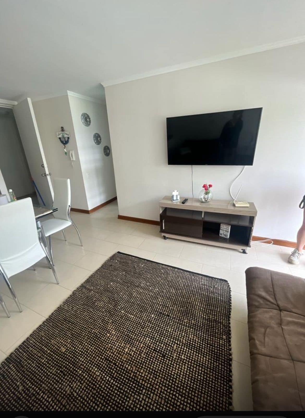 A modern living area features a flat-screen television mounted on a light-colored wall, with a small wooden entertainment unit below. A dark area rug contrasts the tile flooring, while a white dining table with four chairs is positioned nearby. Natural light illuminates the space.