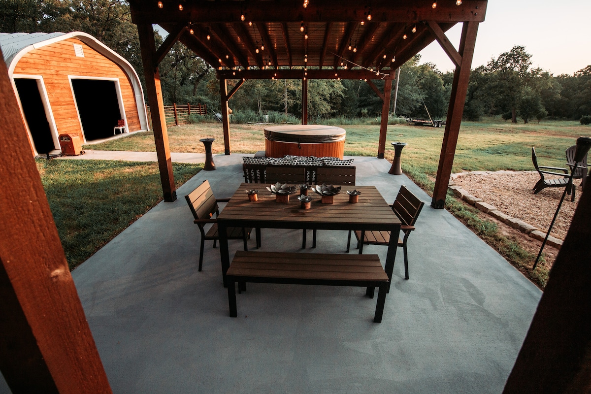 An outdoor seating area features a spacious table with benches under a wooden pergola adorned with string lights. A hot tub is positioned nearby, with open grassy space extending in the background, creating a relaxed setting for gatherings.