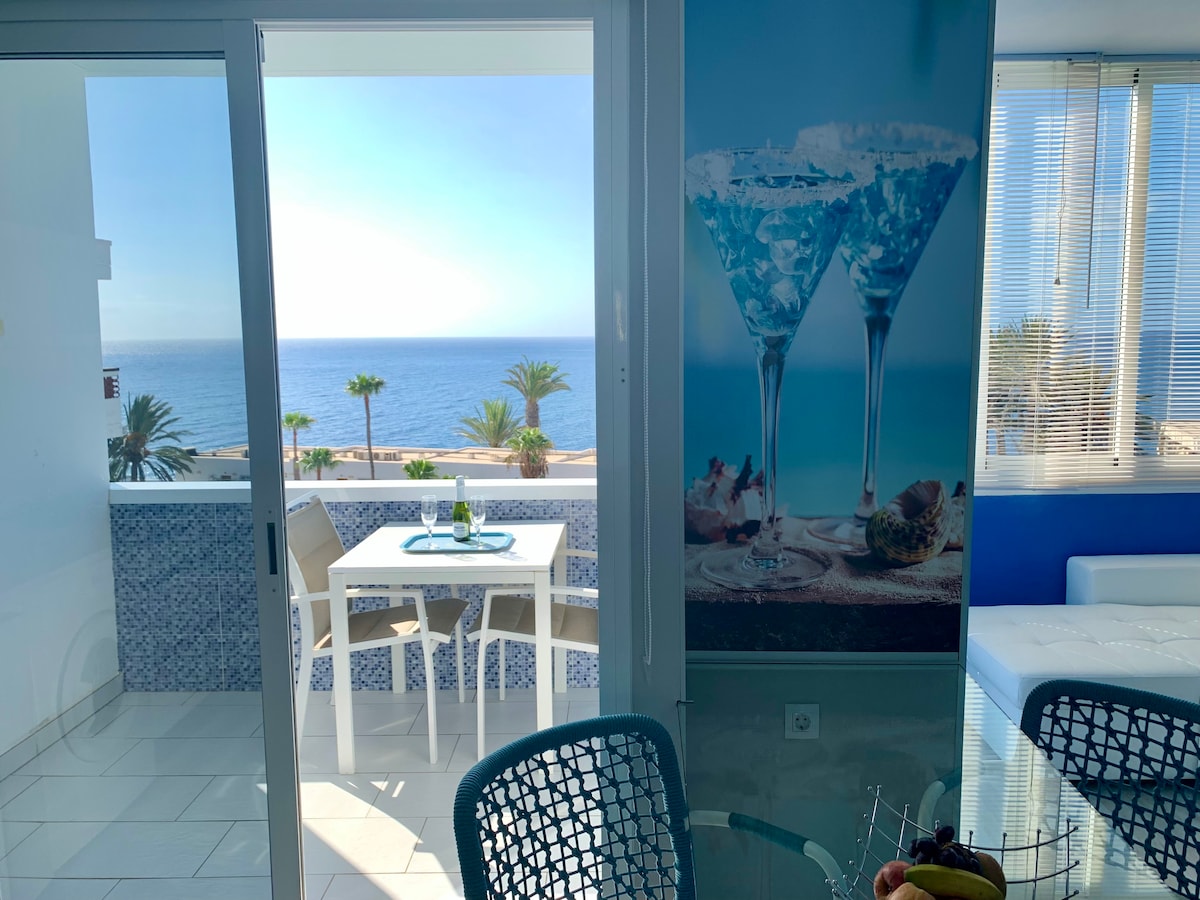 A bright indoor area is visible, featuring a dining table set for two. Large glass doors frame the stunning ocean view, with palm trees in the background. The blue accent wall and decor enhance the serene atmosphere, blending indoor and outdoor living.