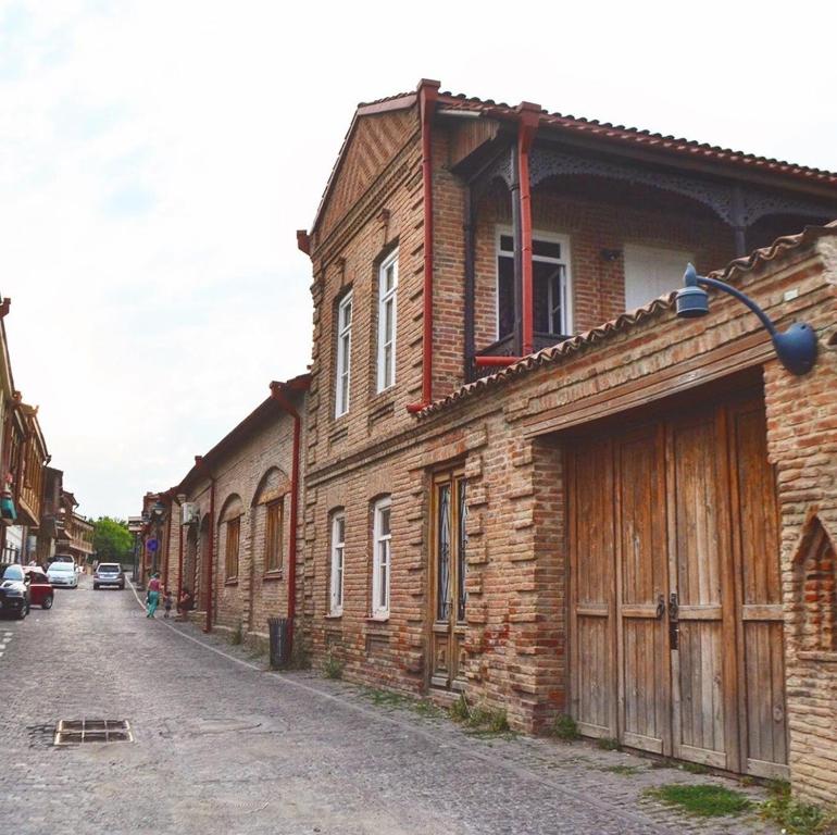 A street scene showcases historic brick buildings, reflecting traditional architectural styles. A cobblestone path extends along the structures, which feature wooden doors and window shutters. Sparse greenery can be seen along the edges, contributing to the serene atmosphere of the surroundings.