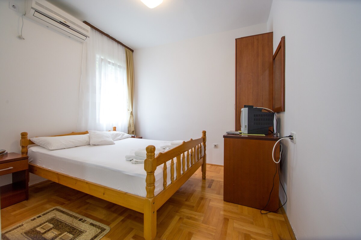 A bright room features a wooden double bed with fresh linens, complemented by a wardrobe. A small desk holds a television and a mini-fridge. The warm wooden flooring adds to the cozy ambiance, while natural light enters through the window adorned with sheer curtains.