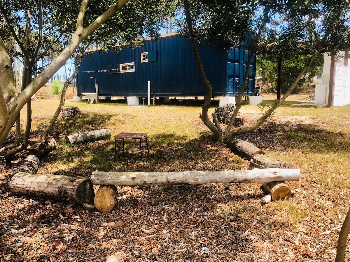 A spacious outdoor area is framed by trees, featuring a wooden log arranged as a bench. A small metal table is positioned nearby, with a large blue container home visible in the background. The grounds are covered in natural foliage and offer a peaceful environment.