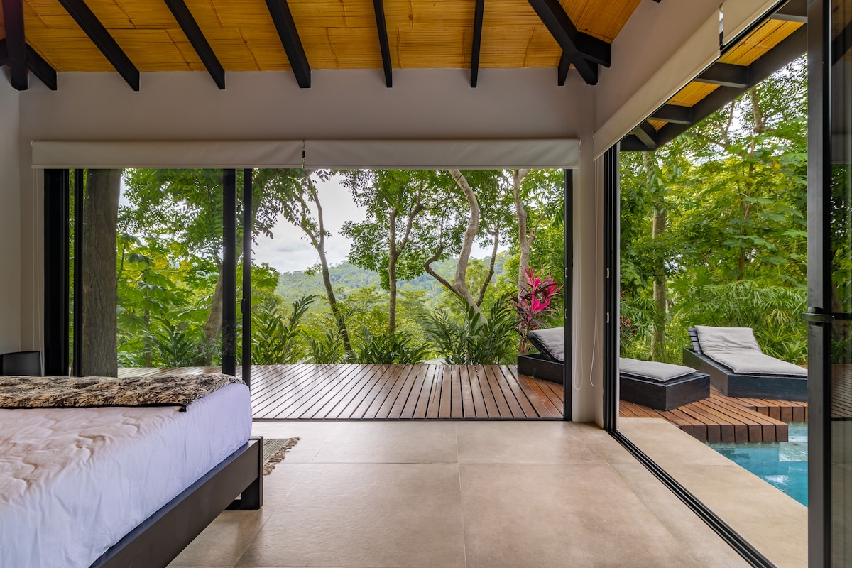 A spacious bedroom features large glass doors that open to a wooden deck surrounded by lush greenery. Sunlight enters through the opening, highlighting a comfortable bed and offering views of the trees and landscape beyond.